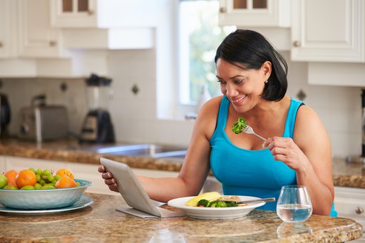 Photo of Black woman eating broccoli at kitchen counter while looking at tablet